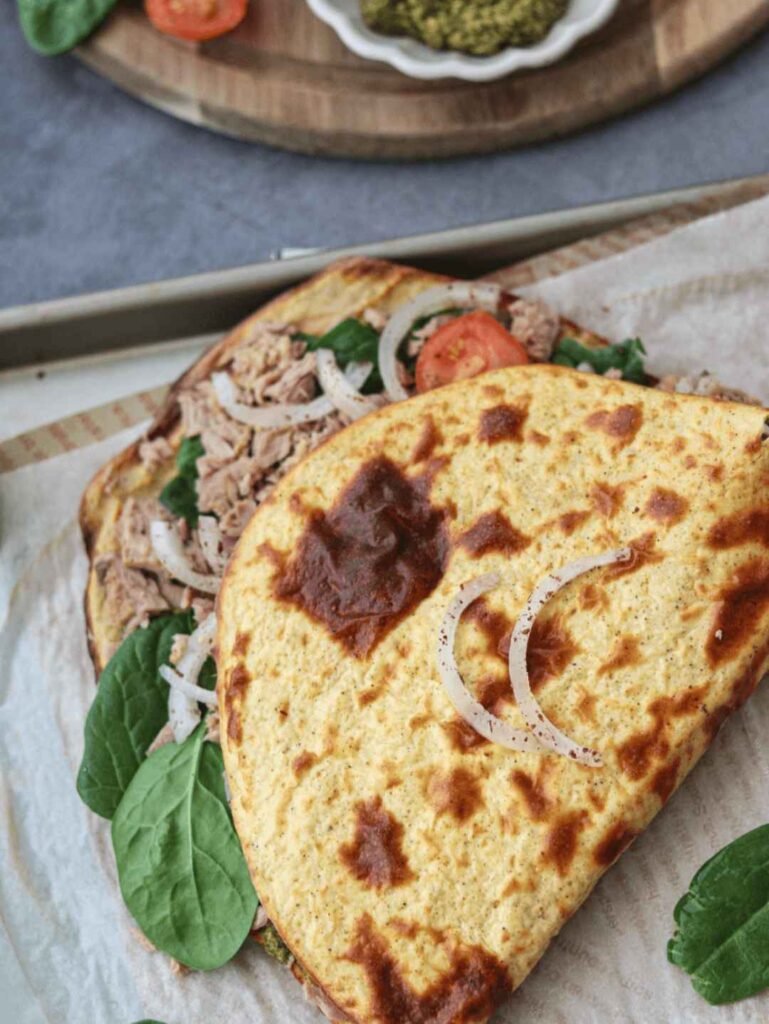 Close-up of a crispy golden flatbread sandwich filled with shredded tuna, fresh spinach leaves, tomato slices, and onion rings, served on parchment paper with pesto sauce in the background.